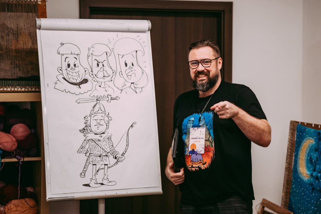 Maciej Kisiel, rysownik i twórca komiksów, uśmiecha się i wskazuje na odbiorcę, stojąc obok tablicy z rysunkami postaci komiksowych